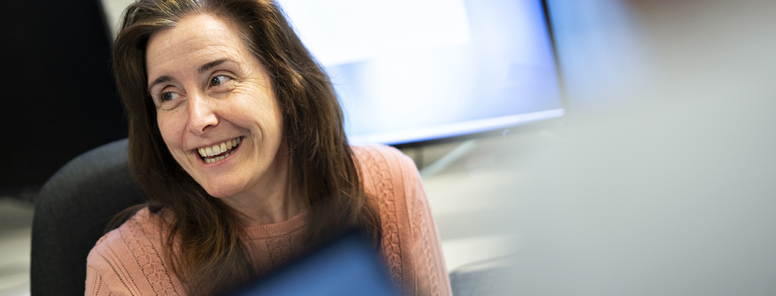 Dark-haired woman sitting in front of computer screens, in pink shirt, with a smile on her face.