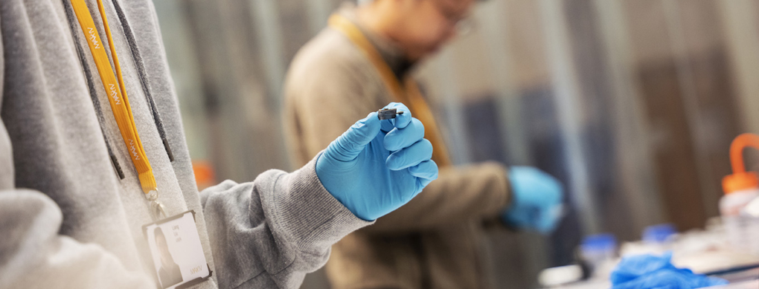 Two young men in a lab wearing light blue plastic gloves studying small parts in engineering research
