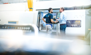 Two men from company Seco Tools standing in front of metal tool cutting machine in working clothes, seen from behind.