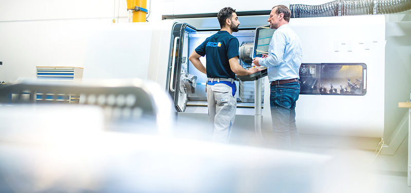 Two men from company Seco Tools standing in front of metal tool cutting machine in working clothes, seen from behind.