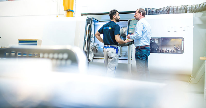 Two men from company Seco Tools standing in front of metal tool cutting machine in working clothes, seen from behind.