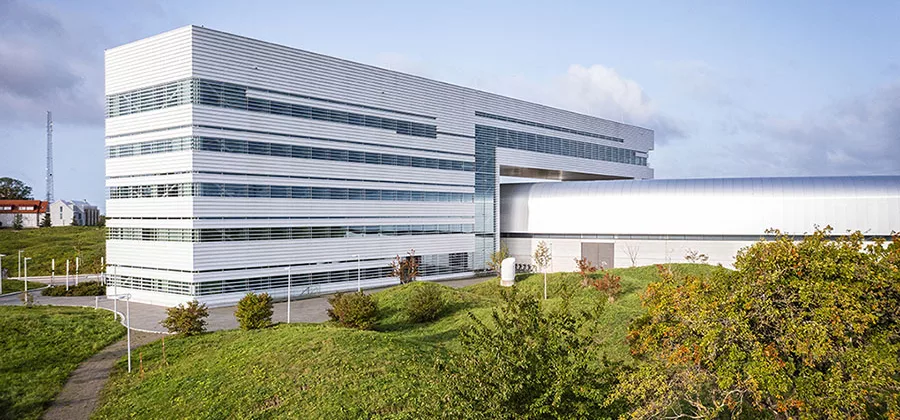 Image of the external MAX IV building with a focus on the office building, trees and green hills in the surroundings.